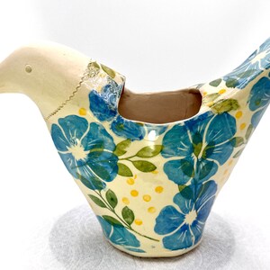 May include: A ceramic bird-shaped planter with a cream-colored head and a brown beak. The body features a pattern of blue flowers, green leaves, and small yellow dots on a cream background. The interior of the bird-shaped vessel is brown.