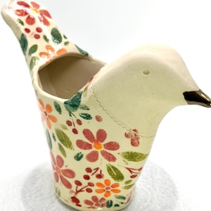 May include: A ceramic bird-shaped vessel with a cream-colored base, featuring a colorful floral pattern of red and orange flowers, green leaves, and small red berries. The bird has a smooth, unpainted head and a dark brown pointed beak.