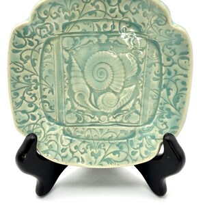 Puede incluir: Un plato cuadrado de cerámica con esquinas redondeadas, con un esmalte azul claro sobre una base crema. El centro muestra un relieve de dos conchas en espiral, mientras que el borde está decorado con un intrincado patrón floral.