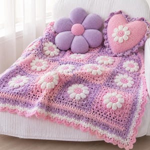 May include: A crochet blanket featuring a pattern of white daisy motifs on a background of purple and pink squares, finished with a scalloped edge. A purple flower-shaped cushion and a pink heart-shaped cushion with a white daisy sit on top.