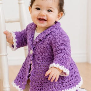 May include: A long-sleeved purple crochet cardigan featuring a collared neckline, a single front button closure, and a flared hem. The garment is detailed with white scalloped trim along the cuffs, the collar, and the bottom edge of the jacket.