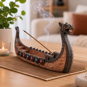 Könnte beinhalten: Ein brauner und dunkelgrauer Räucherstäbchenhalter aus Kunstharz in Form eines Wikingerschiffs mit Drachenkopf-Bug. Das Schiff ist an den Seiten mit Reihen kleiner runder Schilde in Rot und Blau verziert, aus denen ein Räucherstäbchen Rauch aufsteigen lässt.
