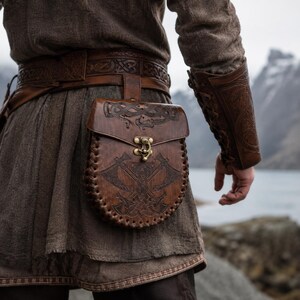 Può includere: Una cintura in pelle marrone con intricati disegni intagliati e una borsa abbinata. La borsa presenta un lembo decorativo con una chiusura e un'immagine dettagliata di asce incrociate. L'outfit include una protezione per il braccio abbinata.