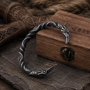 Può includere: Un bracciale a fascia in metallo color argento caratterizzato da una texture intrecciata simile a una corda lungo tutta la banda. Le due estremità del bracciale sono modellate come teste di animali stilizzate, con un piccolo dettaglio circolare.