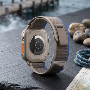 May include: A close-up of a smartwatch with a tan and blue fabric band. The watch case is a light metallic color with an orange button. The watch is resting on a dark gray surface near rocks and a map.