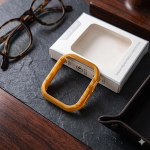 Pode incluir: Uma moldura quadrada de madeira clara para caixa de relógio está sobre uma superfície escura e texturizada, ao lado de uma caixa branca com a etiqueta "WATCH CASE". A moldura tem um padrão de grão de madeira natural. A caixa diz "SHOCK-PROOF PERFECT PROTECTION".
