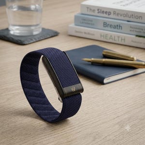 May include: A navy blue fabric wristband featuring a textured weave pattern and a dark wooden clasp component. The clasp is engraved with a small white logo consisting of two vertical lines and a V shape. The band has a flexible, circular design.