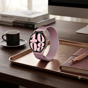 Puede incluir: Un reloj inteligente con esfera negra redonda que muestra la hora 10:08 en grandes números rosas. El reloj cuenta con una caja de metal en oro rosa y una correa de malla metálica rosa a juego. El dispositivo está colocado sobre una bandeja.