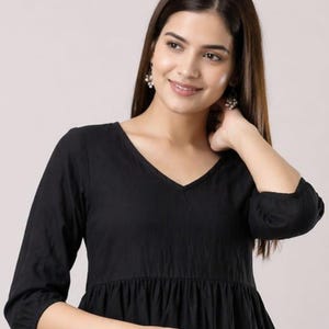 May include: Black V-neck dress with three-quarter sleeves. The dress features a gathered waist and a flowing skirt. The fabric looks to be a lightweight material. The model is wearing pearl earrings.