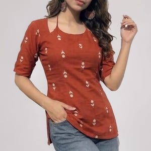 May include: A rust-colored short-sleeved top featuring a white floral pattern, a square neckline with thin halter straps, and side slits, paired with blue denim jeans. The garment includes a small white rectangular label reading Double-tap to zoom.