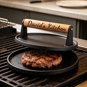 Puede incluir: Una prensa para hamburguesas de hierro fundido con un mango de madera, estampada con "David's Kitchen". La prensa está sobre una hamburguesa cocinada en una parrilla. La prensa es negra y el mango es marrón claro.