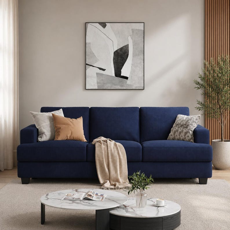 May include: A navy blue three-cushion sofa with a textured fabric. Decorative pillows in neutral tones and a beige throw blanket adorn the sofa. A round coffee table and abstract art complete the living room setting.