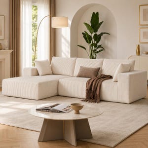 May include: A cream-colored sectional sofa with a corduroy texture, featuring a chaise lounge. A brown throw blanket and decorative pillows add warmth. A round coffee table with a marble top and a beige base sits in front of the sofa.