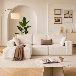 May include: A cream-colored sectional sofa with a corduroy texture, paired with a brown throw blanket and accent pillows. A round coffee table with a book and a bowl sits on a neutral rug. A gold floor lamp and wall art complete the living room setting.