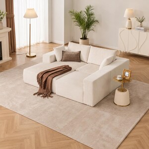 May include: A cream-colored sectional sofa with a chaise lounge, accented by throw pillows and a brown fringed blanket. The sofa sits on a neutral-toned rug, with a gold floor lamp and side table nearby. A potted plant and decorative cabinet complete the living room setting.