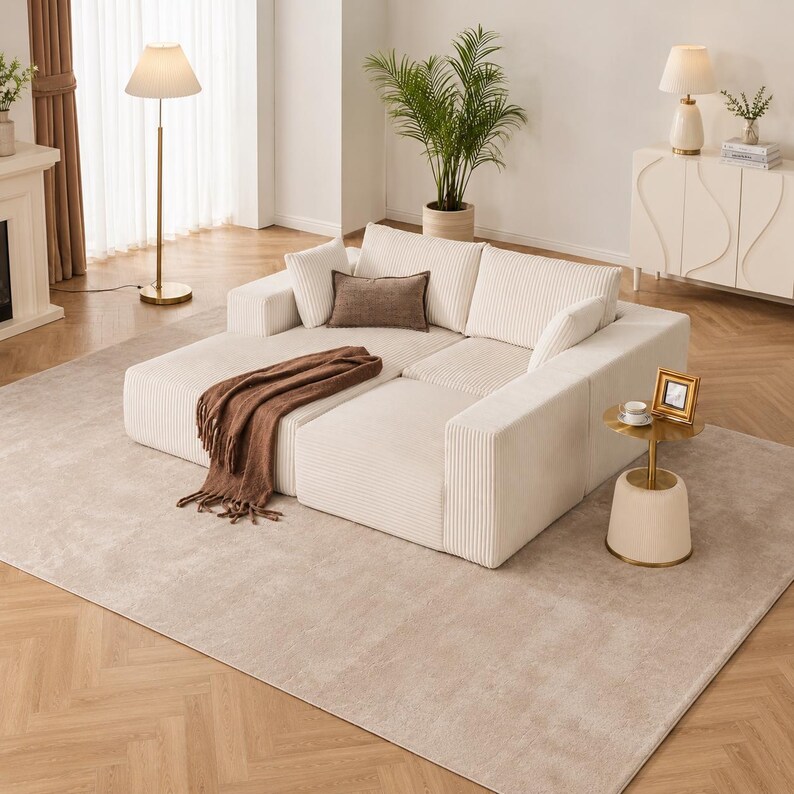 May include: A cream-colored sectional sofa with a chaise lounge, accented by throw pillows and a brown fringed blanket. The sofa sits on a neutral-toned rug, with a gold floor lamp and side table nearby. A potted plant and decorative cabinet complete the living room setting.