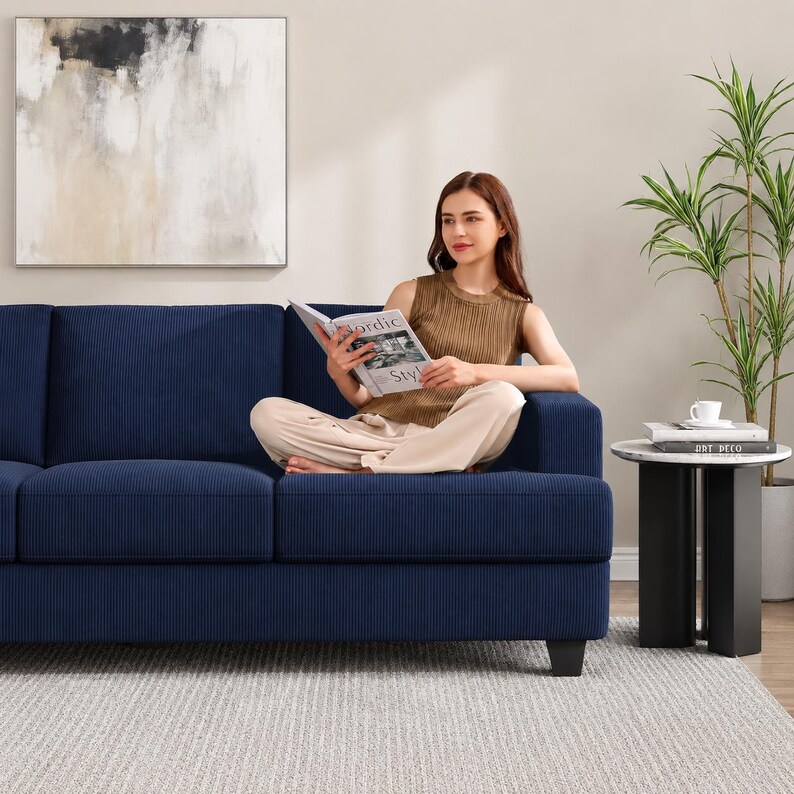 May include: A navy blue corduroy sofa with a person sitting on it, reading a book. The sofa has a modern design with a low profile and square arms. A small side table with a marble top and black legs is next to the sofa.