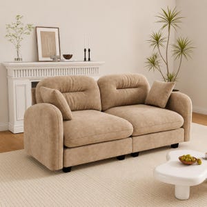 May include: A two-seat beige sofa with rounded arms and a tufted back. The sofa has a matching throw pillow and sits on black feet. The sofa is in a living room setting with a white fireplace and a white coffee table.