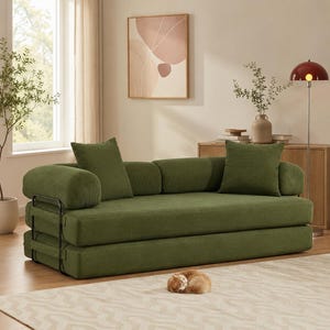May include: A green sofa with a modern design. The sofa has a low profile and features a corduroy-like texture. It includes two square pillows and two bolster pillows. A black metal frame supports the base. A small, curled-up cat rests on the rug in front of the sofa.