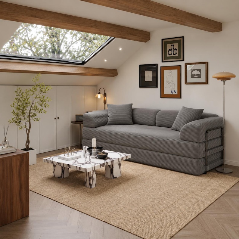 Puede incluir: Un sal&oacute;n moderno con un sof&aacute; de pana gris, obras de arte enmarcadas y una mesa de centro de m&aacute;rmol. La habitaci&oacute;n presenta una paleta de colores neutros con una alfombra beige, vigas de madera y una claraboya, creando un ambiente luminoso y espacioso.