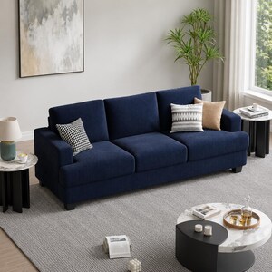 May include: A navy blue three-cushion sofa with a corduroy texture. The sofa has a low profile and is accessorized with decorative throw pillows in various patterns and colors. The sofa is placed on a gray rug, with two side tables and a coffee table.