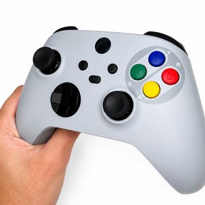 May include: A light grey games controller with black joysticks, buttons, and a directional pad. The controller features coloured buttons: blue, green, red, and yellow. The controller is held in a person's hand against a white background.