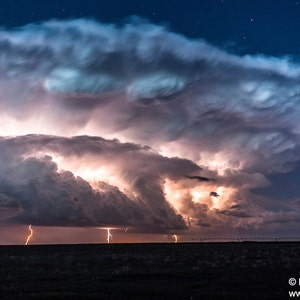 May include: A dramatic night sky with a large, dark storm cloud illuminated by lightning strikes. The cloud has a swirling, textured appearance, and the lightning bolts are bright and jagged. The sky is a deep blue, and there are a few stars visible.