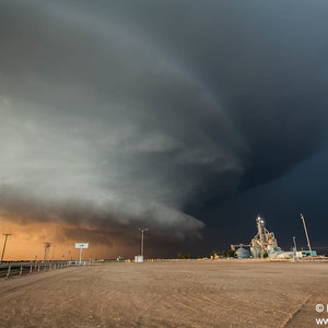 May include: A large, dark storm cloud hangs low over a rural landscape, casting a dramatic shadow over a grain elevator and surrounding fields. The sky is a mix of dark gray and light blue, with a hint of orange in the distance.
