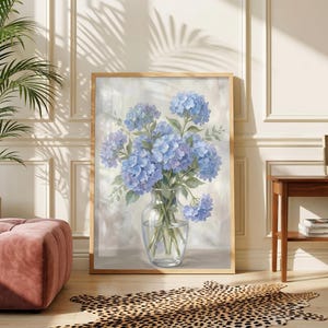 Pode incluir: Impressão emoldurada de flores de hortênsia azuis num vaso de vidro transparente. A obra de arte é exibida numa sala com um pufe rosa, um tapete com estampa de leopardo e uma mesa lateral de madeira. A moldura é de madeira clara.