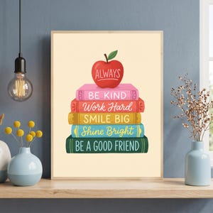 May include: A rectangular wall art print featuring a stack of five colorful books topped with a red apple. The book spines display the text: ALWAYS, BE KIND, Work Hard, SMILE BIG, Shine Bright, and BE A GOOD FRIEND in various fonts and colors.