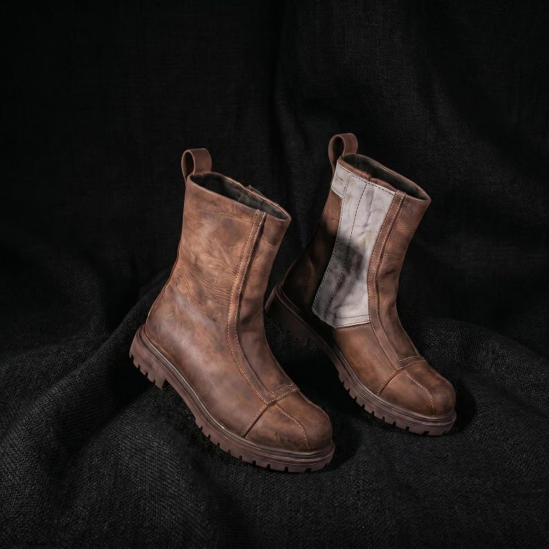 May include: A pair of mid-calf boots crafted from distressed brown leather. The boots feature a rounded toe, a thick lugged rubber sole, and a vertical panel of lighter grey fabric along the outer side. Each boot includes a pull tab at the top.