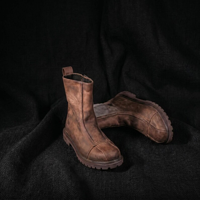 May include: A pair of mid-calf brown leather boots featuring a rounded toe, a vertical seam running up the front, a pull tab at the top, and a thick, lugged rubber sole. The leather has a distressed, matte finish with visible tonal variations.