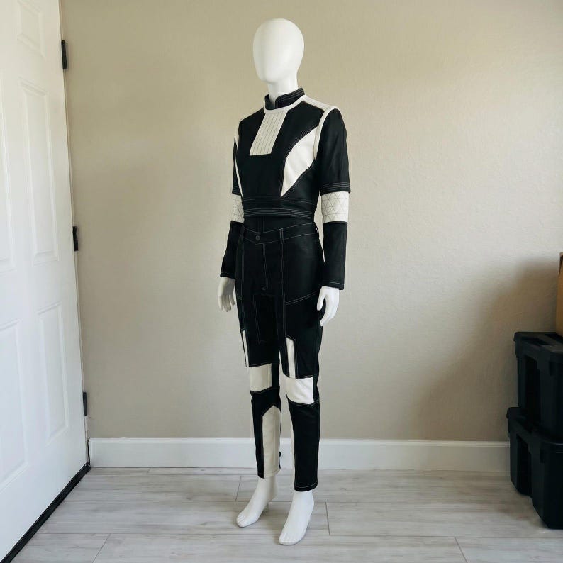 May include: A two-piece outfit consisting of a black and white long-sleeve top with a high collar and matching black and white trousers. The top features a pleated chest panel and white geometric accents. The trousers have white panels on the legs.