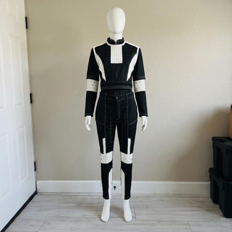 May include: A two-piece outfit consisting of a black long-sleeve top and matching pants with white geometric paneling. The top features a high collar and a pleated chest detail. The pants have white accents on the thighs and lower legs.