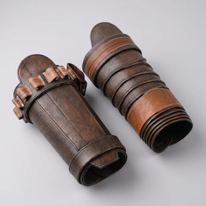 Peut inclure: Une paire de prot&egrave;ge-bras marron fonc&eacute; avec une finition textur&eacute;e. L'un des prot&egrave;ge-bras comporte une s&eacute;rie de petites boucles sur le bord sup&eacute;rieur, tandis que l'autre pr&eacute;sente un design enveloppant avec une bande marron clair lisse.