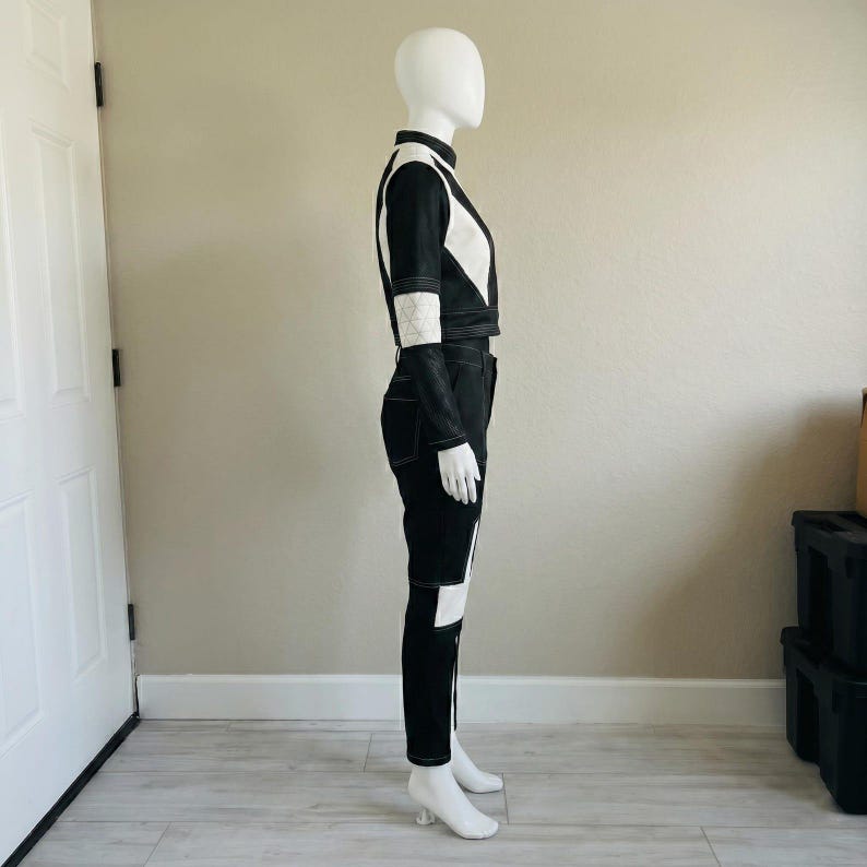 May include: A two-piece outfit consisting of a long-sleeved cropped jacket and matching trousers. The set is primarily black with white color-blocked panels and prominent white topstitching throughout, featuring a quilted texture on the sleeves.