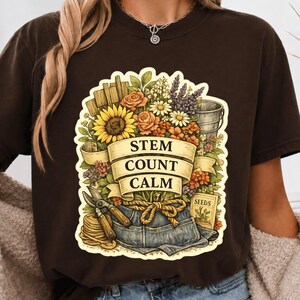 Peut inclure: Un t-shirt marron foncé avec une impression graphique représentant une scène de jardin avec des tournesols, des roses, de la lavande, un seau en métal, des sécateurs, une corde et un sachet de graines. Un ruban affiche STEM COUNT CALM.