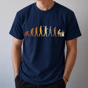 以下が含まれることがあります： ネイビーブルーの半袖Tシャツ。霊長類から始まり、様々な段階を経てコンピュータデスクに向かって座る人物に至るまでの、人間の進化の過程を描いたマルチカラーのグラフィックプリントが施されています。