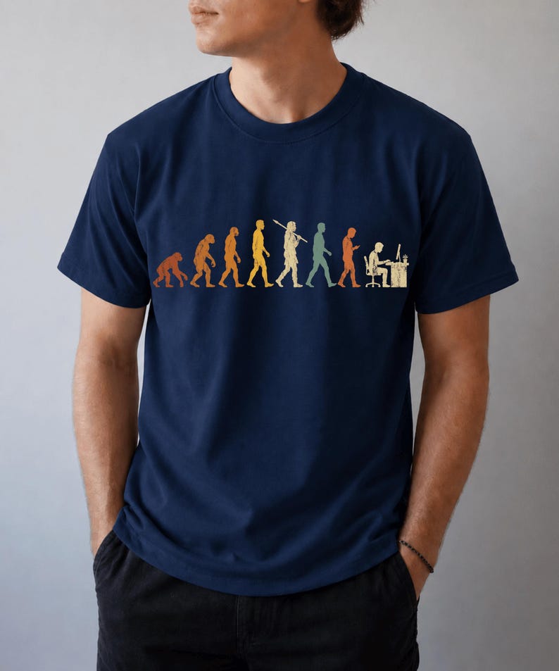 以下が含まれることがあります： ネイビーブルーの半袖Tシャツ。霊長類から始まり、様々な段階を経てコンピュータデスクに向かって座る人物に至るまでの、人間の進化の過程を描いたマルチカラーのグラフィックプリントが施されています。