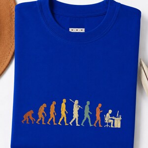 以下が含まれることがあります： ロイヤルブルーのクルーネックTシャツ。人間の進化を描いたマルチカラーのグラフィックプリントが施されています。デザインは、霊長類からコンピュータデスクに座る現代人への進化の過程を表しています。M-M-Mと書かれた白いタグが付いています。