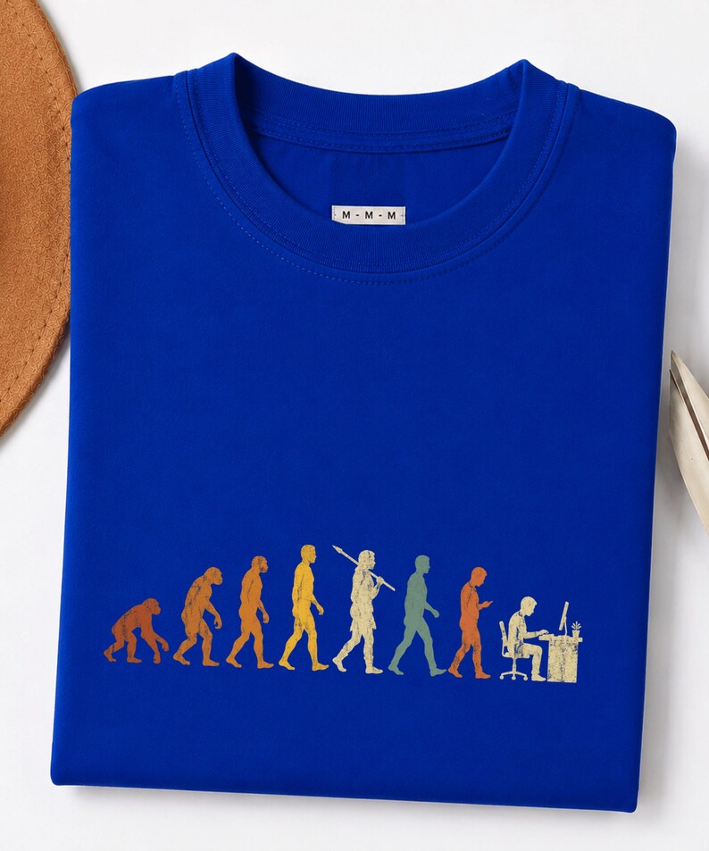 以下が含まれることがあります： ロイヤルブルーのクルーネックTシャツ。人間の進化を描いたマルチカラーのグラフィックプリントが施されています。デザインは、霊長類からコンピュータデスクに座る現代人への進化の過程を表しています。M-M-Mと書かれた白いタグが付いています。