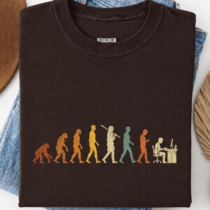 以下が含まれることがあります： ダークブラウンのクルーネックTシャツ。霊長類から始まり、様々な段階を経て、モニターのあるコンピュータデスクに座っている人物へと至る、人類の進化を描いたマルチカラーのグラフィックプリントが施されています。