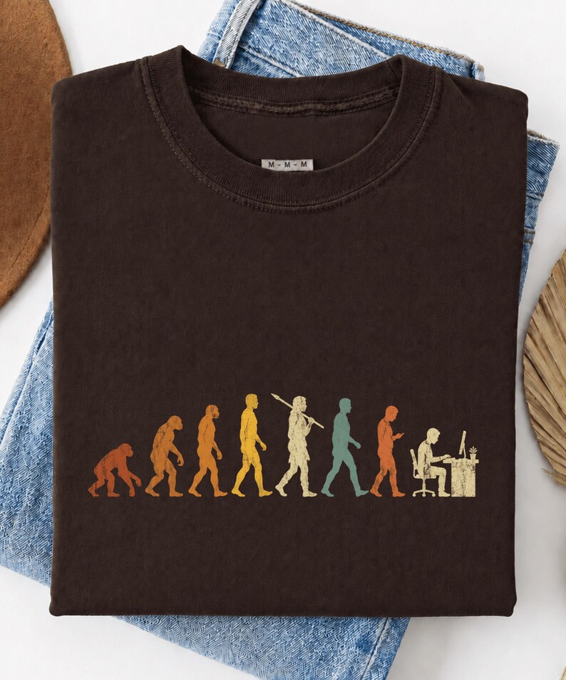 以下が含まれることがあります： ダークブラウンのクルーネックTシャツ。霊長類から始まり、様々な段階を経て、モニターのあるコンピュータデスクに座っている人物へと至る、人類の進化を描いたマルチカラーのグラフィックプリントが施されています。