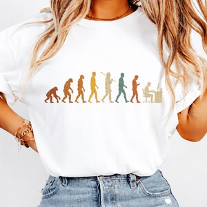 以下が含まれることがあります： 白い半袖Tシャツ。霊長類から始まり、様々な段階を経てデスクでコンピュータに向かって作業する人物に至るまでの人類の進化を描いた、マルチカラーのグラフィックプリントが施されています。