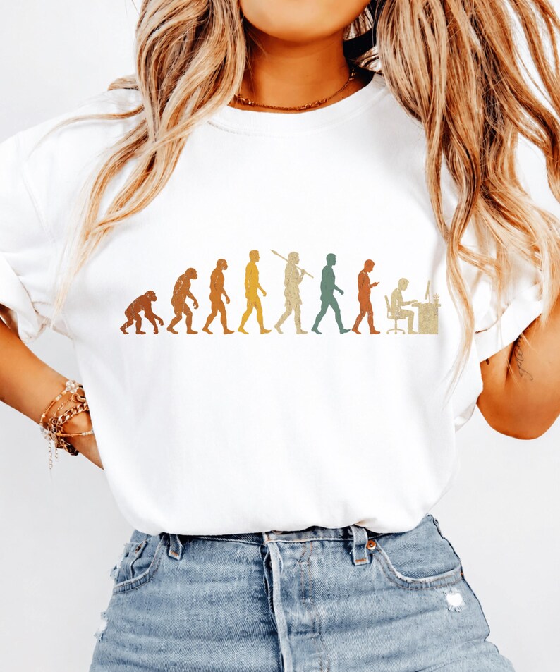 以下が含まれることがあります： 白い半袖Tシャツ。霊長類から始まり、様々な段階を経てデスクでコンピュータに向かって作業する人物に至るまでの人類の進化を描いた、マルチカラーのグラフィックプリントが施されています。