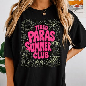 Peut inclure: Un t-shirt noir avec un motif graphique portant le texte TIRED PARAS SUMMER CLUB en rose vif. Le design comprend des illustrations d'un soleil, d'un palmier, d'un parasol, d'un sac fourre-tout, d'une fleur d'hibiscus, de tongs et d'une chaise longue.