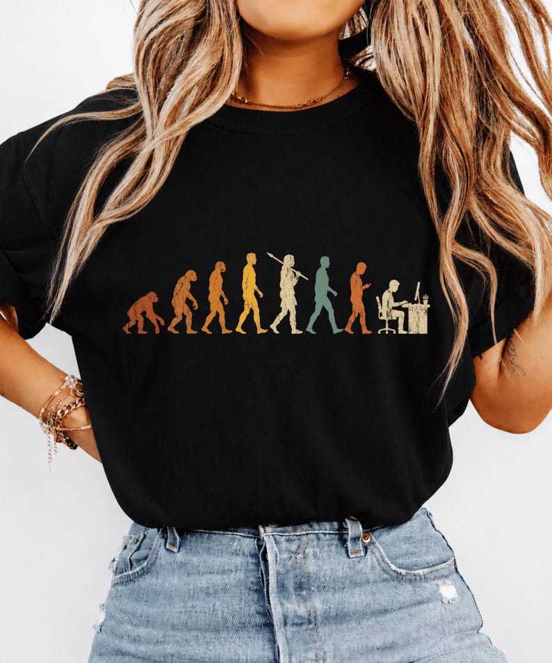 以下が含まれることがあります： 黒いTシャツで、霊長類から始まり、様々な段階を経てデスクでコンピュータに向かって作業する人物に至るまでの、人間の進化の過程を描いたマルチカラーのグラフィックプリントが施されています。