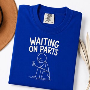 Peut inclure: Un t-shirt bleu roi avec un motif blanc représentant un bonhomme allumette assis sur un seau et tenant un boulon. Le texte au-dessus du personnage indique "WAITING ON PARTS" dans une police blanche grasse. Le t-shirt porte une étiquette Comfort Colors.