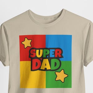 May include: A beige t-shirt featuring a colorful graphic that reads "SUPER DAD." The graphic is divided into four squares of red, blue, yellow, and green, with yellow stars in the corners. The text is in black with a yellow outline.