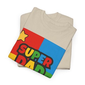 May include: Two beige t-shirts are folded, with one on top featuring a colorful graphic. The graphic includes a yellow star and the words "SUPER DAD" in bold, multi-colored letters against a red, blue, and yellow background. The shirts are made of a soft material.